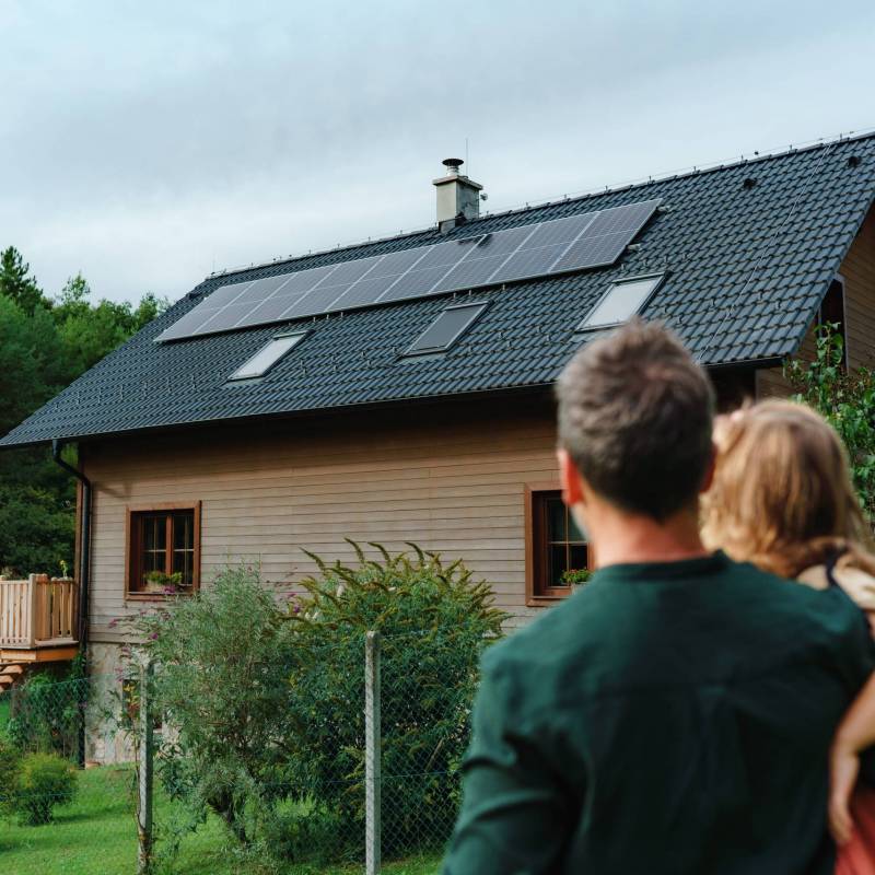 Rear view of dad holding her little girl in arms and looking at their house with solar panels.Alternative energy, saving resources and sustainable lifestyle concept.